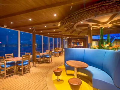 Innenansicht einer geräumigen Bar-Lounge auf der Carnival Vista, mit Holzböden und Decken, sowie großen Fenstern, die auf das dunkle Meer blicken. Blaue Sitzbänke und Holztische mit blauen Stühlen sind vorhanden. Eine große, mit Stroh gedeckte Bar-Struktur und eine Bepflanzung mit Kakteen und orangefarbenen Blumen schafft eine tropische Atmosphäre.