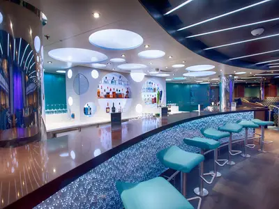 Eine moderne Bar-Lounge auf der Carnival Vista. Die geschwungene Bar hat eine glänzende Oberfläche und eine beleuchtete Vorderseite mit kleinen blauen Lichtpunkten. Türkisfarbene Barhocker stehen davor. Im Hintergrund sind runde Regale mit Flaschen und weitere Sitzbereiche zu sehen.