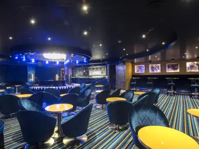 Innenansicht einer stilvollen Bar-Lounge auf der Carnival Vista. Blaue Samtsessel, gelbe Tische, gestreifter Teppich, ein Piano und eine Bar mit blauer Beleuchtung sind zu sehen. Die Decke ist dunkel mit vielen kleinen Lichtern, die einen Sternenhimmel imitieren.