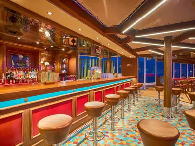 Eine stilvolle Schiffsbar mit langem Holztresen und vielen Barhockern. Der bunte Mosaikboden und die leuchtende blaue Lichtleiste unterstreichen das moderne Design. Durch die Fenster ist die blaue Nacht zu sehen.