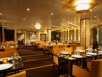 Ein luxuriöses Restaurant an Bord der Carnival Vista, festlich gedeckt mit hellen Stühlen und glänzenden Tischen. Warme goldene Beleuchtung und Kunstwerke schmücken den Raum.