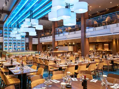 Ein modernes, helles Restaurant auf der Carnival Vista mit zahlreichen gedeckten Tischen. Im Hintergrund eine beleuchtete Bar und eine obere Ebene mit weiteren Sitzplätzen. Blaue Lichtakzente an Decke und Bar.