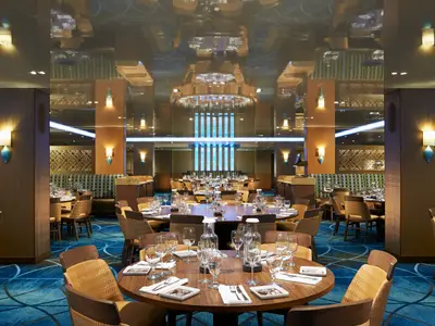 Elegantes, modernes Restaurant auf der Carnival Vista. Gedeckte Holztische und Stühle, blaue Lichtsäulen und spiegelnde Decke. Warme Wandleuchten und blauer Teppich. Ohne Personen.
