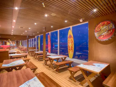 Helles Burger-Restaurant im Surfer-Stil auf der Carnival Vista. Holztische, Bänke und bunte Surfbretter an großen Fenstern. Rechts ein großes 'Guy's Burger Joint' Logo.