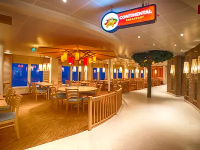 Ein hell beleuchtetes, modernes Restaurant an Bord der Carnival Vista, mit Holztischen, gepolsterten Stühlen und großen Fenstern. Ein Schild weist auf ein kontinentales Frühstück hin. Pflanzen und eine Ziegelwand schmücken den Raum.