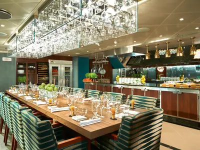 Innenansicht eines eleganten Spezialitätenrestaurants an Bord. Eine lange Holztisch ist festlich gedeckt, umgeben von gepolsterten Stühlen mit blau-grünen Mustern. Im Hintergrund ist eine offene Küche mit Metalloberflächen, hängenden Töpfen und frischen Zutaten sichtbar. Über dem Tisch hängen gläserne Beleuchtungskörper mit vielen Weingläsern.