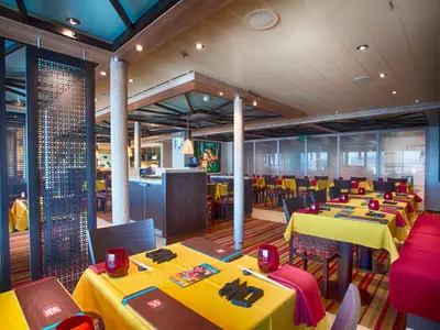 Innenansicht eines farbenfrohen Spezialitätenrestaurants an Bord der Carnival Vista. Gelb gedeckte Tische mit rotem Geschirr und Stühlen. Dekorative Raumteiler mit geometrischem Muster. An den Wänden asiatische Kunst.