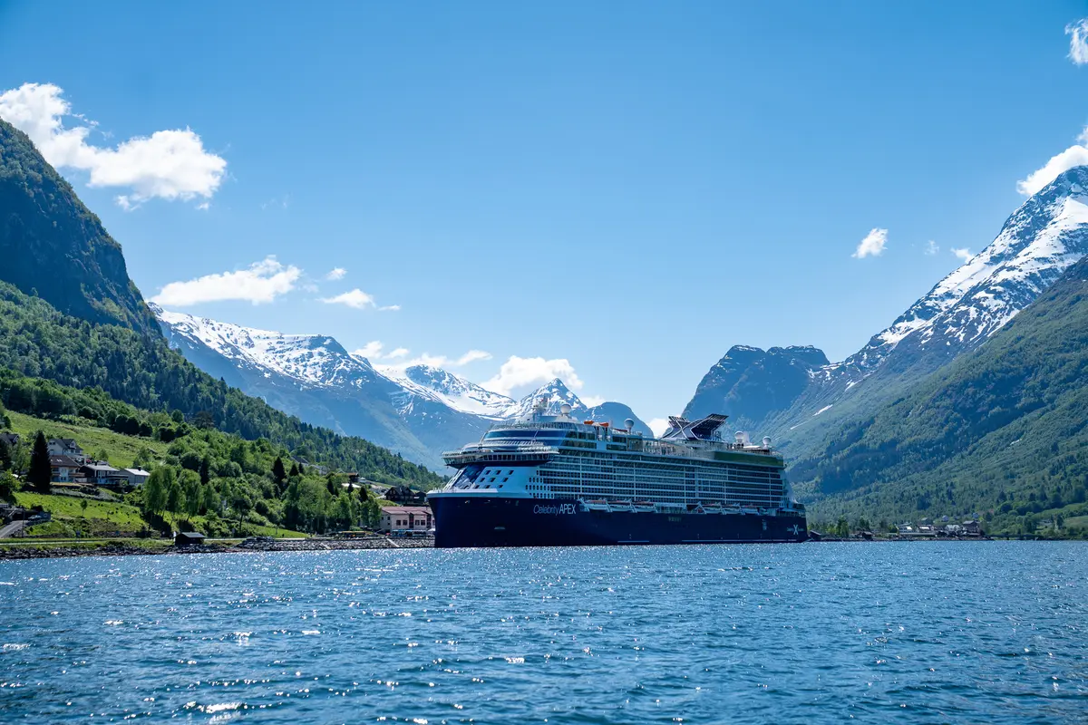 Ein grosses, dunkelblaues Kreuzfahrtschiff mit dem Namen Celebrity Apex liegt auf ruhigem Wasser in einem Fjord, umgeben von grünen und schneebedeckten Bergen unter blauem Himmel mit wenigen Wolken.