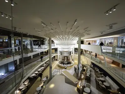 Blick ins moderne Atrium der Celebrity Apex. Zentrale Bar unter auffälliger Deckenbeleuchtung. Mehrere Ebenen mit Glasbalustraden und Sitzbereichen, belebt von wenigen Personen.