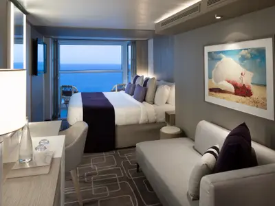 Moderne Kabine der Celebrity Apex: Doppelbett, Sofa, Schreibtisch. Bodentiefe Fenster zeigen Meer und Balkon. Kunstwerk an der Wand. Helle Einrichtung.