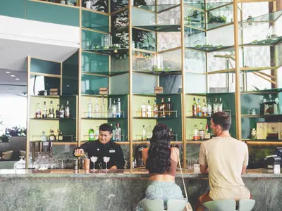 Ein Barkeeper im schwarzen Hemd mixt an einer Bar mit Marmorplatte Cocktails. Im Hintergrund Regale mit Flaschen und Dekoration. Zwei Gaeste stehen mit dem Ruecken zur Kamera an der Bar.