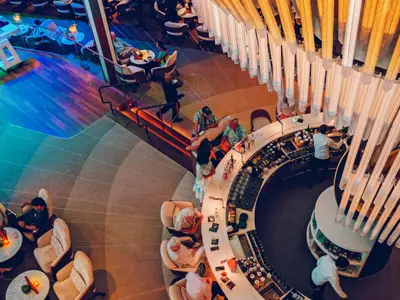 Blick von oben auf einen mehrstufigen Barbereich an Bord der Celebrity Apex. Gäste entspannen in Loungesesseln und an der runden Bar, beleuchtet von einer modernen Deckeninstallation.