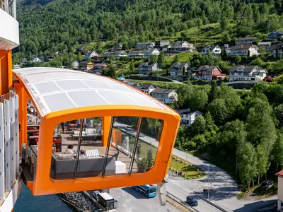 Der orangefarbene Magic Carpet der Celebrity Apex mit sitzenden Gästen. Im Hintergrund sind grüne Hügel mit Häusern, eine Straße und ein Bus zu sehen.