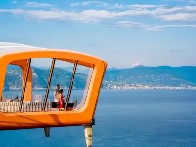 Ein Paar steht im orangefarbenen Magic Carpet der Celebrity Apex, einer gläsernen Plattform über dem Meer, und blickt auf die bergige Küste mit einer Stadt. Blauer Himmel.