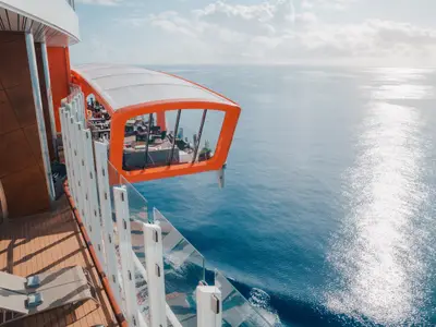 Orangefarbener Magic Carpet der Celebrity Apex, eine Plattform mit Sitzbereichen, schwebt über blauem Meer. Schiffsdeck mit Sonnenliegen und Glasgeländern im Vordergrund.