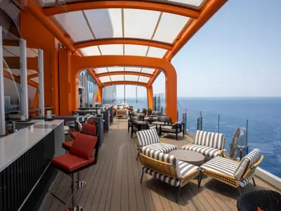 Ein modernes Außendeck der Celebrity Apex mit einer Bar auf der linken Seite und roten Barhockern. Mehrere Lounge-Sitzgruppen mit gestreiften Polstern und kleinen Tischen sind unter einem markanten orangefarbenen Dach angeordnet. Im Hintergrund erstreckt sich der weite Ozean unter blauem Himmel.