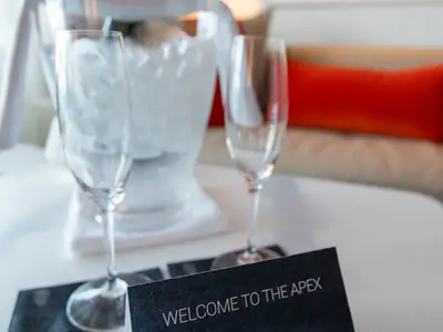 Nahaufnahme von zwei leeren Champagnergläsern und einem Eiskübel auf einem weißen Tisch. Im Hintergrund ist eine orangefarbene Kissenlehne sichtbar.