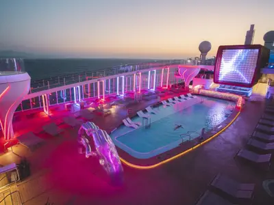 Das beleuchtete Deck der Celebrity Apex zeigt abends einen Pool mit Schwimmer und Liegestühlen. Bunte Neonlichter, eine Skulptur und ein großer Bildschirm prägen die Szene mit Meerblick.