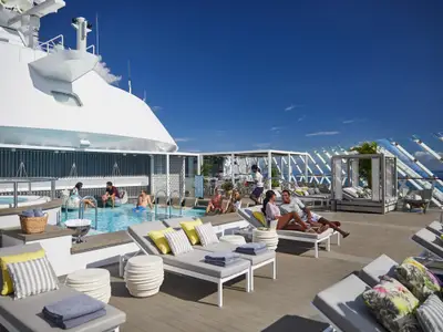 Panoramablick auf das Sonnendeck der Celebrity Apex. Gäste genießen den Pool, Jacuzzi und moderne Lounge-Bereiche unter strahlend blauem Himmel. Eine Bedienung liefert Getränke an die Sonnenliegen.