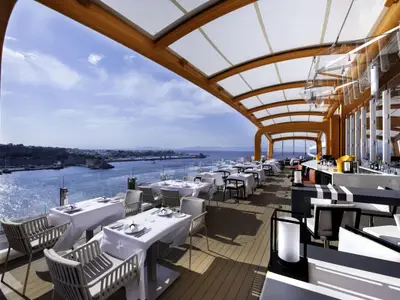 Ein modernes Außendeck-Restaurant des Kreuzfahrtschiffs Celebrity Apex mit gedeckten Tischen, Bar und Meerblick. Im Hintergrund ist eine sonnige Hafenstadt zu erkennen.