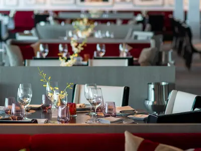 Elegant gedeckter Tisch im Restaurant der Celebrity Apex. Gläser, gelbe Orchideen und rote Kerzenhalter sind zu sehen. Im unscharfen Hintergrund weitere Tische und Essbereiche.