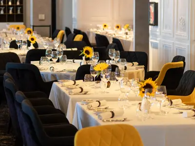 Ein elegantes Restaurant mit vielen runden Tischen, die festlich mit weissen Tischdecken, Geschirr, Gläsern und kleinen Sonnenblumen-Arrangements gedeckt sind. Die Stühle sind dunkel, einige haben gelbe Akzente.