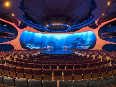 Theater auf der Celebrity Apex mit Rundbühne, geschwungenen Sitzen. Große LED-Wand zeigt Unterwasserwelt mit Mantarochen, blau-orange beleuchtet.