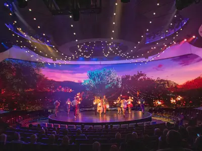 Blick auf eine Theaterbühne, auf der mehrere Darsteller in bunten Kostümen tanzen und Laternen tragen. Im Hintergrund eine große Videoleinwand, die eine Landschaft bei Sonnenuntergang mit Bäumen und Lichtern zeigt. Das Publikum ist im Vordergrund zu sehen.