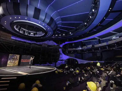 Innenansicht eines modernen Theaters oder Konferenzraums mit geschwungener blauer Deckenbeleuchtung. Eine Person spricht auf einer runden Bühne vor drei Bildschirmen. Im Publikum tragen viele Menschen gelbe Schutzhelme.