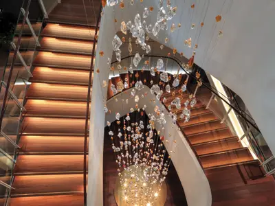 Blick von oben auf eine breite Holztreppe mit Stufenbeleuchtung und einem Geländer aus Glas und Holz. Über der Treppe hängt eine opulente Lichtinstallation aus Hunderten von glitzernden, unregelmäßig geformten Kristallen in Weiß und Bernstein, die von der Decke herabhängen. Das Atrium ist in warmen Brauntönen und Weiß gehalten.