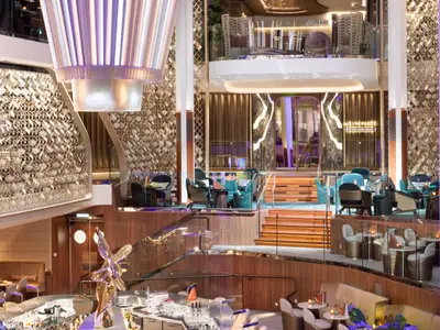 Das elegante, mehrstöckige Atrium der Celebrity Ascent. Es zeigt moderne Sitzgruppen, Glasgeländer, Holztreppen, eine große Leuchte und goldene Wandstrukturen.