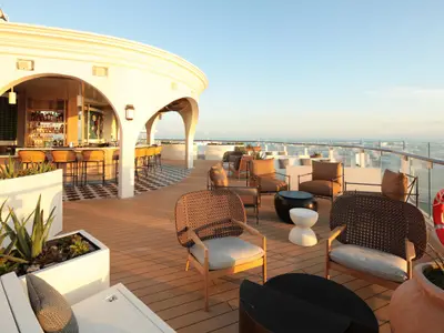 Sonniges Outdoor-Deck der Celebrity Ascent mit Bar, Lounge-Möbeln auf Holzdeck, Pflanzen und Meerblick. Die Bar hat gewebte Hocker. Weites Meer unter blauem Himmel im Hintergrund.