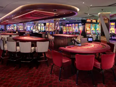 Leeres Casino der Celebrity Ascent. Rote Spieltische und Stühle im Vordergrund. Reihen bunter Spielautomaten im Hintergrund. Roter Teppich und moderne Beleuchtung.