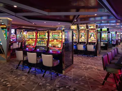 Modernes Casino an Bord der Celebrity Ascent. Reihen leuchtender Spielautomaten mit Stühlen füllen den Raum. Ein Barbereich mit Hockern ist rechts. Eleganter Teppich und gedämpftes Licht dominieren.