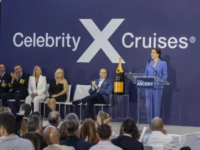 Eine Frau in blauem Anzug spricht an einem Pult mit dem Celebrity Ascent Logo auf einer Bühne. Hinter ihr ist ein "Celebrity X Cruises" Banner und sechs Personen sitzen und applaudieren.