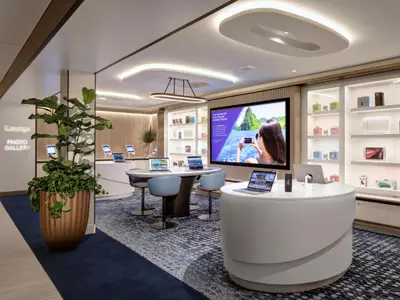 Moderne iLounge auf der Celebrity Ascent: Weiße Tische mit Laptops und Tablets. Großer Bildschirm und beleuchtete Elektronikregale. Eine Zierpflanze steht auf blauem Teppich.