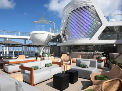Ein sonniges Außendeck der Celebrity Ascent mit modernen Lounge-Möbeln, Sonnenschirmen und einer Bar. Futuristische Schiffarchitektur und eine Stadtkulisse prägen das Bild.