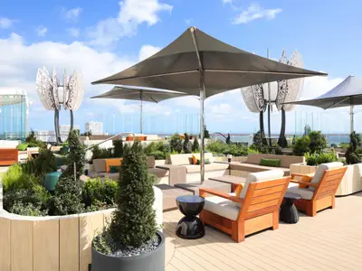 Auf dem Rooftop Garden der Celebrity Ascent laden bequeme Loungemöbel, umgeben von Pflanzen und kleinen Bäumen, zum Entspannen ein. Große Sonnenschirme spenden Schatten, im Hintergrund sind Gebäude und das Meer zu sehen.