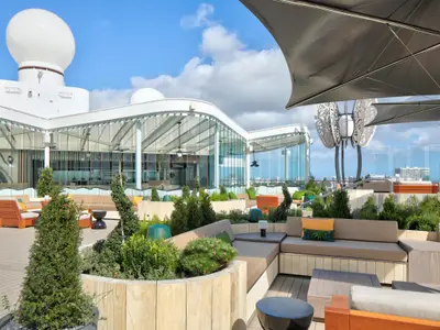 Ansicht des Außendecks der Celebrity Ascent: Moderne Sitzbereiche, Pflanzen, Sonnenschirme. Eine gläserne Lounge und ein weißes Radom im Hintergrund unter blauem Himmel.