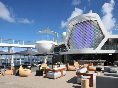Sonniges Außendeck der Celebrity Ascent mit Lounge-Möbeln, Sonnenschirmen und einer Bar. Eine große, farbig leuchtende Wabenwand ist zu sehen, dahinter Meer und Stadtpanorama.