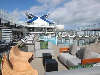 Oberdeck der Celebrity Ascent mit Pool, Liegestühlen, Barhockern an der Poolbar und Lounge-Sitzgruppen. Im Hintergrund die X-förmige Schiffsarchitektur.