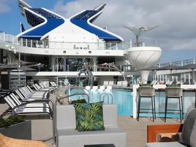 Das Pooldeck der Celebrity Ascent zeigt einen Pool, viele Liegestühle und eine glänzende Oktopus-Skulptur. Auffällige blaue und weiße Schiffsarchitektur und eine Bar sind sichtbar.