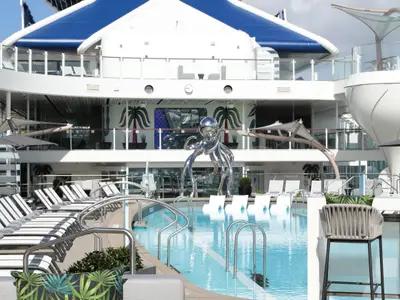 Sonniges Pooldeck der Celebrity Ascent: Blaues Schwimmbecken, umgeben von Sonnenliegen. Eine große, glänzende Oktopus-Skulptur. Im Hintergrund sind weitere Decks des Kreuzfahrtschiffs zu sehen.