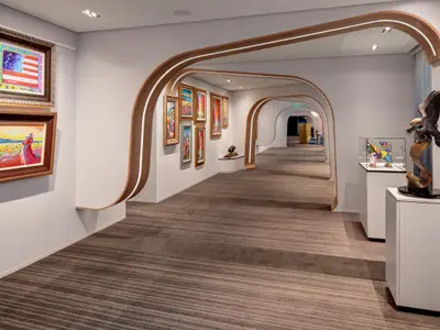 Blick in eine Kunstgalerie an Bord der Celebrity Ascent mit geschwungenen Holzarchitekturelementen. Links hängen zwei Gemälde, rechts stehen Skulpturen und weitere bunte Bilder zieren die Wand.