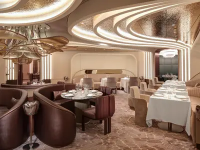 Innenansicht eines luxuriösen Restaurants mit braunen und beigefarbenen Sitzgelegenheiten, runden Tischen mit weißer Tischwäsche und Gläsern, sowie einer auffälligen goldenen Deckenbeleuchtung.