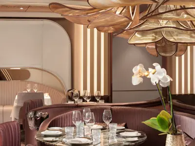Ein elegantes Restaurant mit bequemen, runden Polsterbänken in Brauntönen. Tische sind eingedeckt mit Gläsern. Eine weiße Orchidee steht im Vordergrund. Warme Goldtöne und vertikale Lichtstreifen an der Wand schaffen eine luxuriöse Atmosphäre.