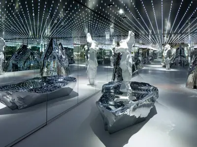 Blick in eine weitläufige, futuristische Lounge, die von Spiegelflächen und unzähligen kleinen Lichtern an der Decke geprägt ist. Im Raum stehen abstrakte, weisse und glänzende silberne Skulpturen, die das Licht reflektieren und eine unendliche Tiefe erzeugen. Der Boden ist hell und spiegelnd, was die Lichtreflexe noch verstärkt. Der Raum wirkt modern und künstlerisch.