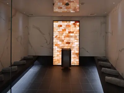 Ein moderner Saunaraum mit einer großen, beleuchteten Wand aus Salzziegeln in der Mitte und dunklen Holzsitzen an den Seiten, auf denen gerollte Handtücher liegen. Der Boden ist mit dunklen Fliesen ausgelegt und die Wände sind mit hellem Marmor verkleidet.