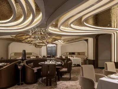 Ein luxuriöses Restaurant mit runden, braunen Ledersofas und Tischen. Die Decke ist goldfarben, wellenförmig und beleuchtet. Eine große, blattförmige Kronleuchter hängt in der Mitte.