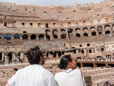 Zwei unscharfe Personen von hinten im Vordergrund, die auf die alte, massive Steinarchitektur des Kolosseums mit mehreren bögenförmigen Ebenen blicken.